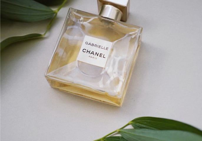 A Chanel Gabrielle bottle laying on a white background with a green foliage surround.